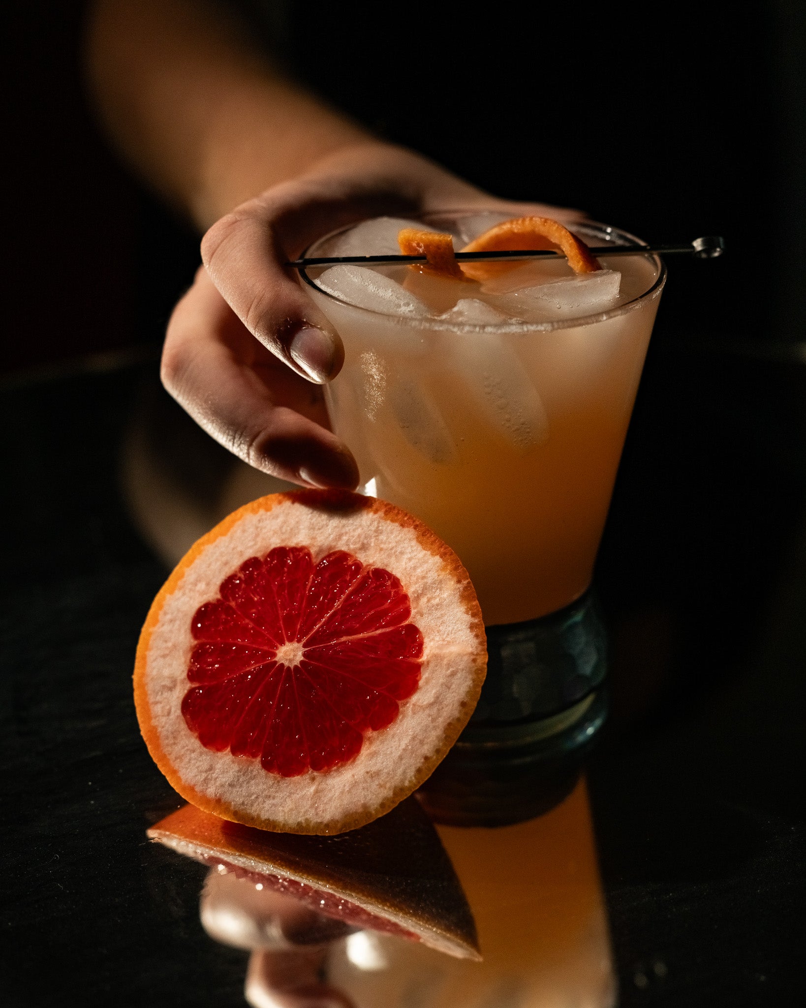 Hand holding a cocktail with ice and an orange slice, garnished with a half-grapefruit, against a dark background.