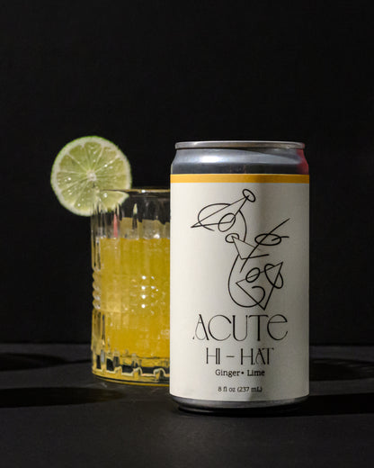 Can of Acute Hi-Hat ginger lime drink with a glass of yellow liquid and lime wheel on a dark background
