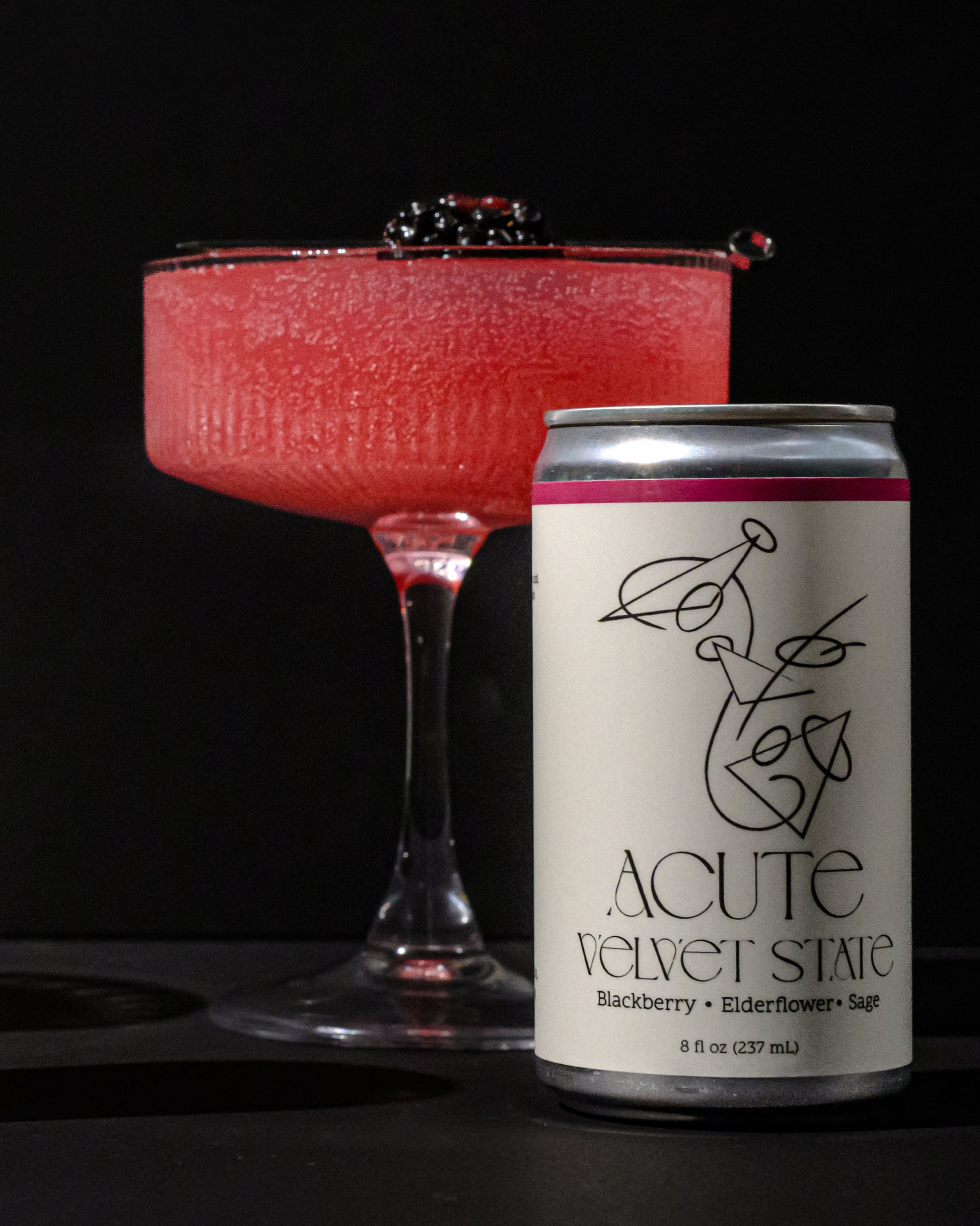 Red cocktail in a glass with a can of 'Acute Velvet State' on a black background